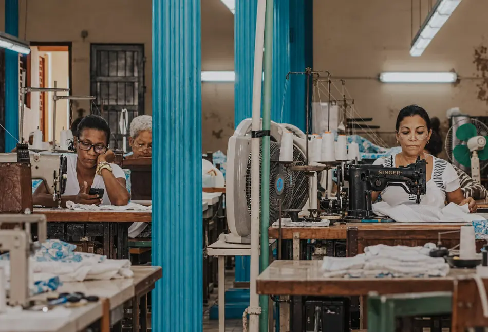 Women working at sewing machines in a Cuban workshop, with turquoise columns and available indoor lighting