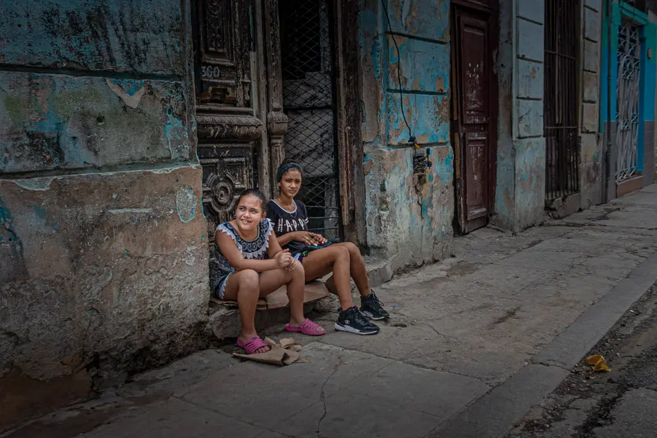 Two girls sitting outside a weathered doorway in Havana, Cuba, photographed by James Nader
