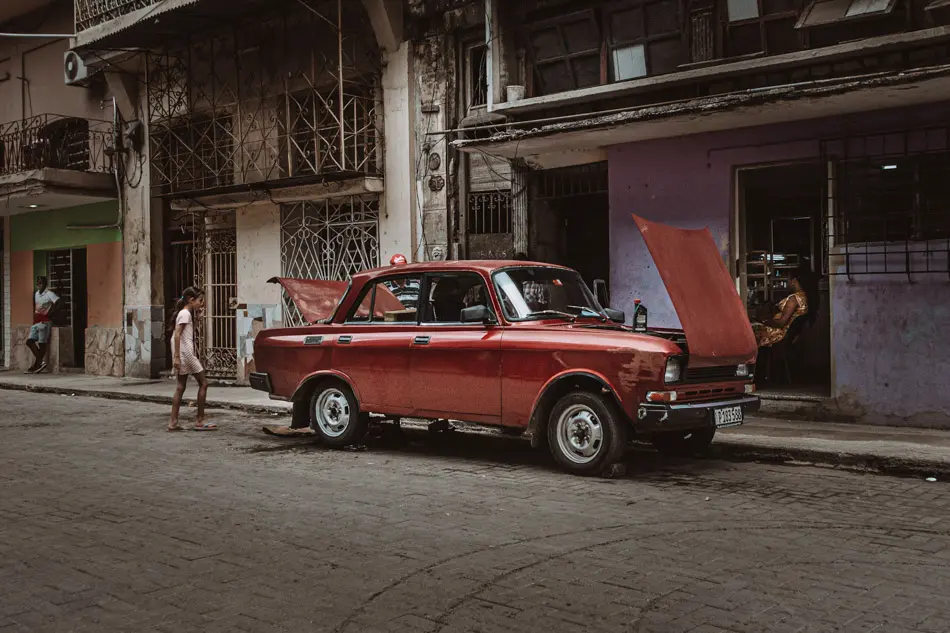 Red vintage car with its bonnet open on a residential street in Havana, with everyday activity in the background